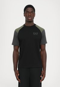 Man standing facing forward wearing black t-shirt with dark gray and green sleeves and EA7 Emporio Armani logo on chest.
