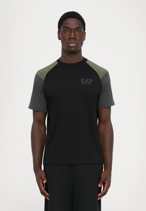 Man standing facing forward wearing black t-shirt with dark gray and green sleeves and EA7 Emporio Armani logo on chest.