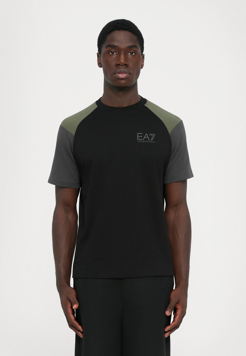 Man standing facing forward wearing black t-shirt with dark gray and green sleeves and EA7 Emporio Armani logo on chest.