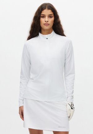 Top blanco de manga larga con cremallera y diseño ajustado, combinado con una falda blanca. La tela tiene una textura suave y presenta detalles de logotipos en las mangas.