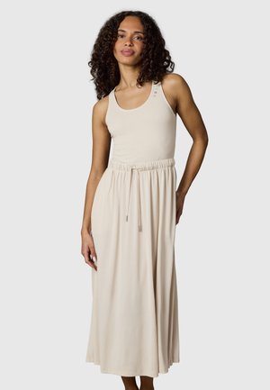 Frau mit lockigem Haar, die ein ärmelloses beiges Maxikleid mit Kordelzug in der Taille und kleinen dekorativen Knöpfen nahe am Ausschnitt trägt.