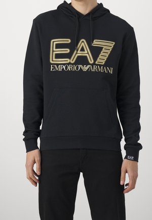 Man draagt een zwarte hoodie met gouden "EA7 Emporio Armani" logo, een voorzak en trekkoorden, gecombineerd met een zwarte broek.