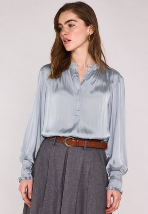 Femme portant un chemisier en satin bleu clair avec des poignets volantés et des boutons, rentré dans une jupe plissée grise taille haute avec une ceinture marron.