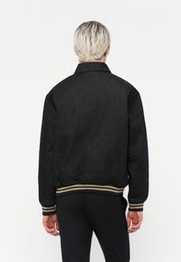 Svart denim bomberjakke med ribbestrikket krage og mansjetter, med et stripet liv. Glatt tekstur, minimalistisk design, ingen synlig merkevare.