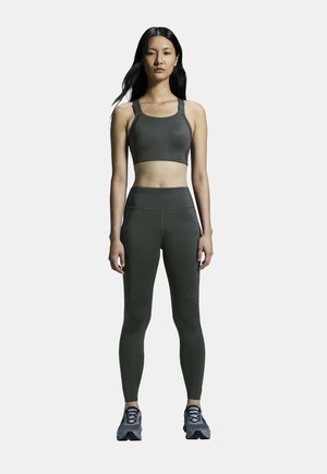 Olivgrüner Sport-BH und hochtaillierte Leggings, die eine glatte Textur und verstärkte Nähte aufweisen. Getragen mit grauen Sportschuhen.
