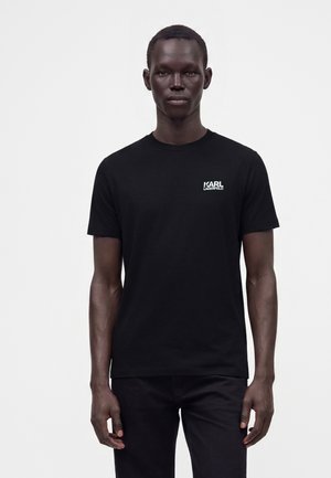 Uomo che indossa una semplice t-shirt nera a maniche corte con il logo "KARL LAGERFELD" sul petto, in piedi davanti a uno sfondo bianco.