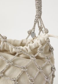Sac en tissu beige avec un design en maille, orné de quincaillerie en ton argent et de détails scintillants. Il présente un haut froncé et une sangle décorative.