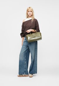 Oliivroheline käekott tähekujulise aksessuaariga, poleeritud tekstuuriga ja kaheastmeliste käepidemetega; mudel kannab pruunikat ülisuurust sviitrit ja laiu teksapükse.