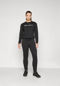Svart hettegenser med "Champion"-logo i hvitt, kombinert med matchende svarte joggebukser. Begge plaggene har en avslappet passform og myk tekstur. Sorte joggesko fullfører antrekket.