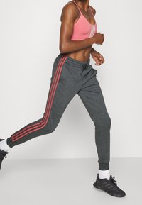 Persona atlética corriendo, vestida con un sujetador deportivo rosa, pantalones deportivos gris oscuro con rayas rosas, calcetines blancos y zapatillas de correr negras.