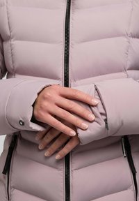 Chaqueta de plumas de color rosa claro con costuras horizontales, cremallera negra y un bolsillo discreto en la manga. Textura suave y aislante.