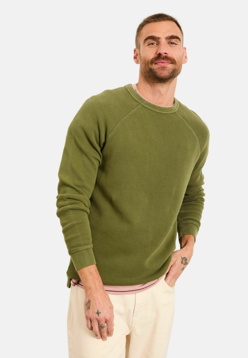 Olivgrüner, gerippter Pullover mit langen Ärmeln und Rundhalsausschnitt, über einem hellrosa Hemd getragen, kombiniert mit cremefarbenen Hosen.