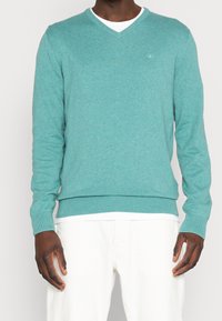 Turkusowy sweter robiony na drutach z dekoltem w serek, długimi rękawami, ściągaczami przy mankietach i u dołu. Posiada małe logo na klatce piersiowej. Stylizowany na białą koszulę.