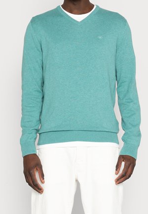 Turkos stickad tröja med V-ringning, långa ärmar, ribbade cuffar och fåll. Har en liten logotyp på bröstet. Styling över en vit skjorta.