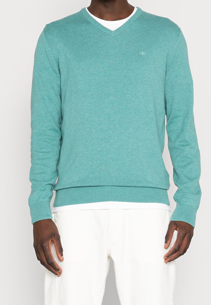 Turkusowy sweter robiony na drutach z dekoltem w serek, długimi rękawami, ściągaczami przy mankietach i u dołu. Posiada małe logo na klatce piersiowej. Stylizowany na białą koszulę.