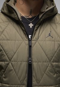 Olivengrön vadderad väst med hög krage, svart dragkedja och Jordan-logotyp. Har en strukturerad tyg och diamantmönster.