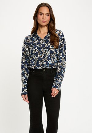 Frau mit langen braunen Haaren, die eine blau-schwarz-weiße langärmelige Bluse mit Blumendruck trägt, die in hoch taillierte schwarze Hosen gesteckt ist, steht vor einem weißen Hintergrund.