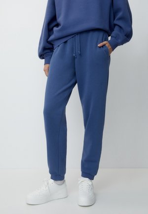 Persoon die blauwe joggerbroek en bijpassend sweatshirt draagt met witte sneakers, staand tegen een effen lichte achtergrond.