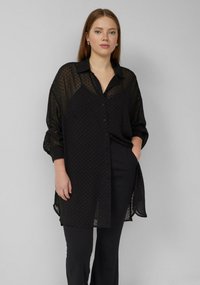 Schwarze transparente Bluse mit Polka-Dot-Muster, Knopfleiste, Kragen und langen Ärmeln. Getragen über schwarze Hosen; leichtes Material.