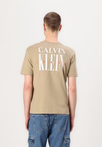 Kaki medvilninis marškinėliai su didelėmis baltomis ir raudonomis raidėmis "CALVIN KLEIN JEANS" nugaroje. Trumpomis rankovėmis, apvaliu kaklu, laisvu siluetu.