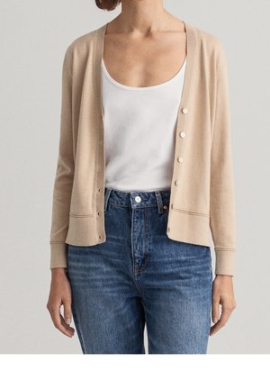 Osoba ubrana w beżowy kardigan zapinany na guziki, na biały top z głębokim dekoltem i niebieskie, wysokim stanem dżinsy, stojąca na jednolitym tle.