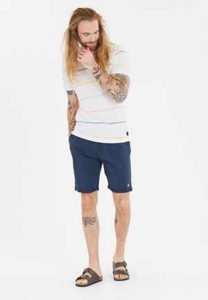 Mænds stribet T-shirt i hvid med blå, pink og gule striber, parret med marineblå shorts og brune sandaler. Bemærkelsesværdige tatoveringer synlige på armene.