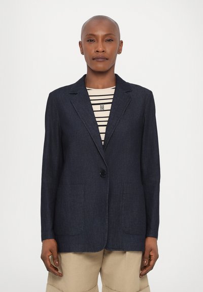 Blazer bleu foncé en tissu tissé, avec un bouton unique, des revers crantés et deux poches patch à l'avant. En dessous, une chemise rayée est visible.