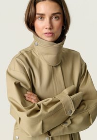 Veste imperméable beige avec un col haut, des manches larges et des accents de boutons-pression. Texture lisse et coupe décontractée.