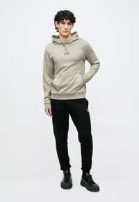 Hele beež kapuutsiga pusa kängurutasku ja paelaga; kombineeritud mustade jooksepükstega. Mõlemal on peened brändingu aktsendid.