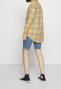 Camicia a quadri gialla con accenti neri e bianchi, abbinata a pantaloni shorts in denim blu. Sneakers alte nere completano l'abito.