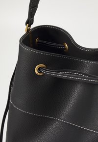 Sac seau en cuir noir texturé avec surpiqûres blanches, œillets dorés et une bandoulière passant par le bord supérieur pour la fermeture.