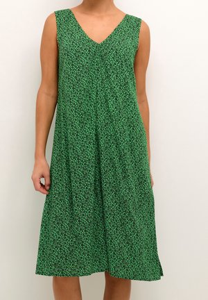 Ärmelloses grünes Midikleid mit kleinem schwarzem Blumenmuster und V-Ausschnitt, getragen von einer stehenden Person vor einfarbigem Hintergrund.