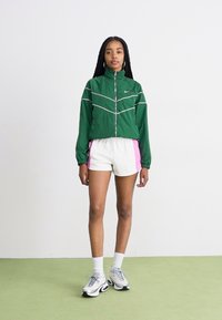 Casaco verde com riscas brancas e fecho éclair, combinado com calções brancos com detalhes em rosa. O modelo usa ténis cinzentos com meias brancas.