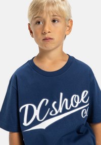 Námořnická modrá bavlněná trička s krátkým rukávem, se značným bílým logem "DC shoe co" tištěným přes přední stranu.