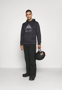 Μαύρο φούτερ με κουκούλα και λογότυπο βουνού, σε συνδυασμό με μαύρο παντελόνι και μποτάκια. Το μοντέλο κρατάει ένα μαύρο κράνος, παρουσιάζοντας μια casual εμφάνιση για εξωτερικούς χώρους.