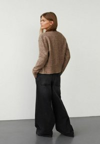 Smeđi pleteni pulover s teksturom u krojenju koji se završava iznad struka, uparen s crnim trapericama širokih nogavica koje imaju stražnje džepove. Model stoji ispred neutralne pozadine.