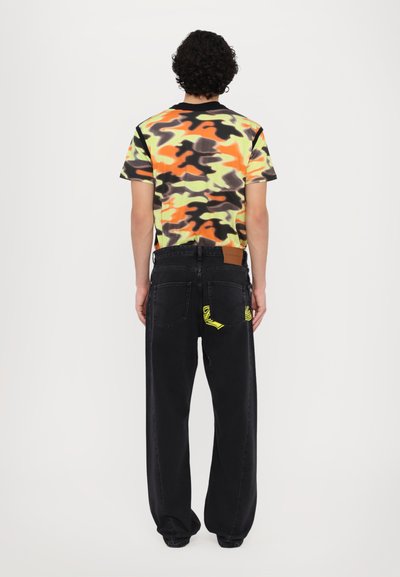 Zwarte denim broek met een losse pasvorm, voorzien van gele accenten en een leren label. Het multicolore camouflage t-shirt heeft korte mouwen.