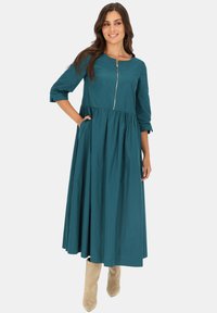 Sukienka w kolorze morskim z długimi rękawami, okrągłym dekoltem, zapinana na zamek z przodu, z marszczoną spódnicą. Posiada boczne kieszenie oraz luźny, swobodny krój.