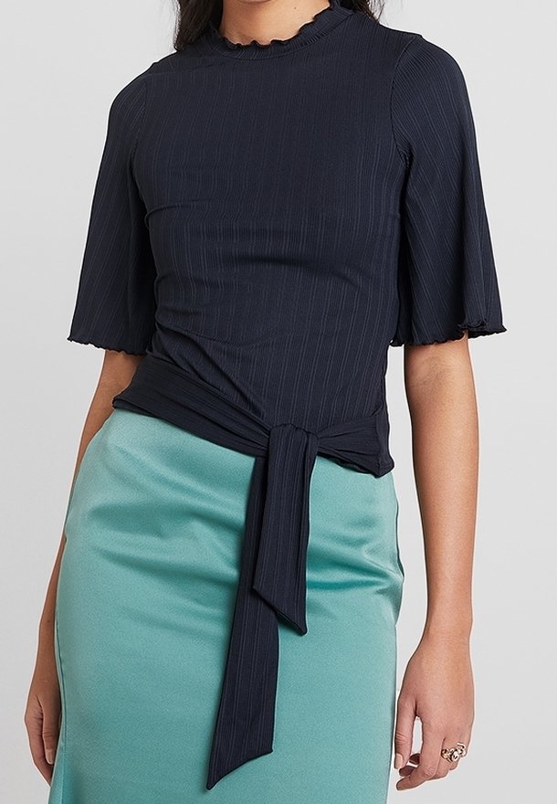 Haut en mailles navy avec des manches courtes évasées et un décolleté festonné. Présente un détail de nœud à l'avant et se marie avec une jupe vert menthe.