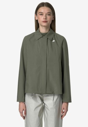 Giovane donna con giacca verde oliva e pantaloni grigio chiaro, in piedi contro uno sfondo chiaro uniforme, vista frontale.