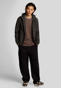 Giacca a zip grigio scuro con cappuccio, t-shirt marrone e pantaloni neri. Dettagli notevoli: logo ricamato e sneakers con motivo.