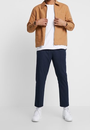 Bruine katoenen jas met rits over een wit shirt, gecombineerd met marineblauwe pantalon met krijtstrepen en witte sneakers. Casual, aansluitend silhouet.