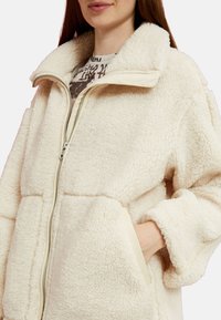 Chaqueta de forro polar color crema con un cuello alto, cierre de cremallera frontal y grandes bolsillos. Presenta una tela suave y texturizada con un ribete en contraste.