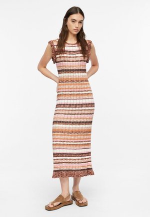 Młoda kobieta stoi z rękami za plecami, ubrana w prążkowaną, paskowaną sukienkę midi i brązowe sandały na platformie na białym tle.