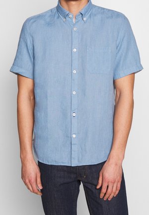 Hombre vistiendo una camisa de manga corta color azul claro con botones y bolsillo en el pecho, y jeans oscuros, mostrado desde los hombros hasta las caderas.