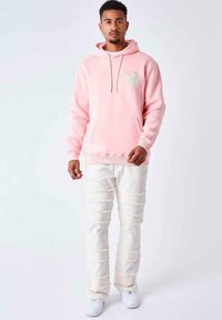 Felpa rosa chiaro con un patch a forma di cuore, colletto con cordino e tasca frontale, abbinata a pantaloni bianchi strappati e sneaker bianche.
