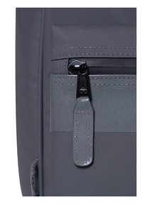 Sezione di uno zaino in tessuto grigio con una tasca con cerniera dotata di un tirante nero e una finitura testurizzata sul materiale.