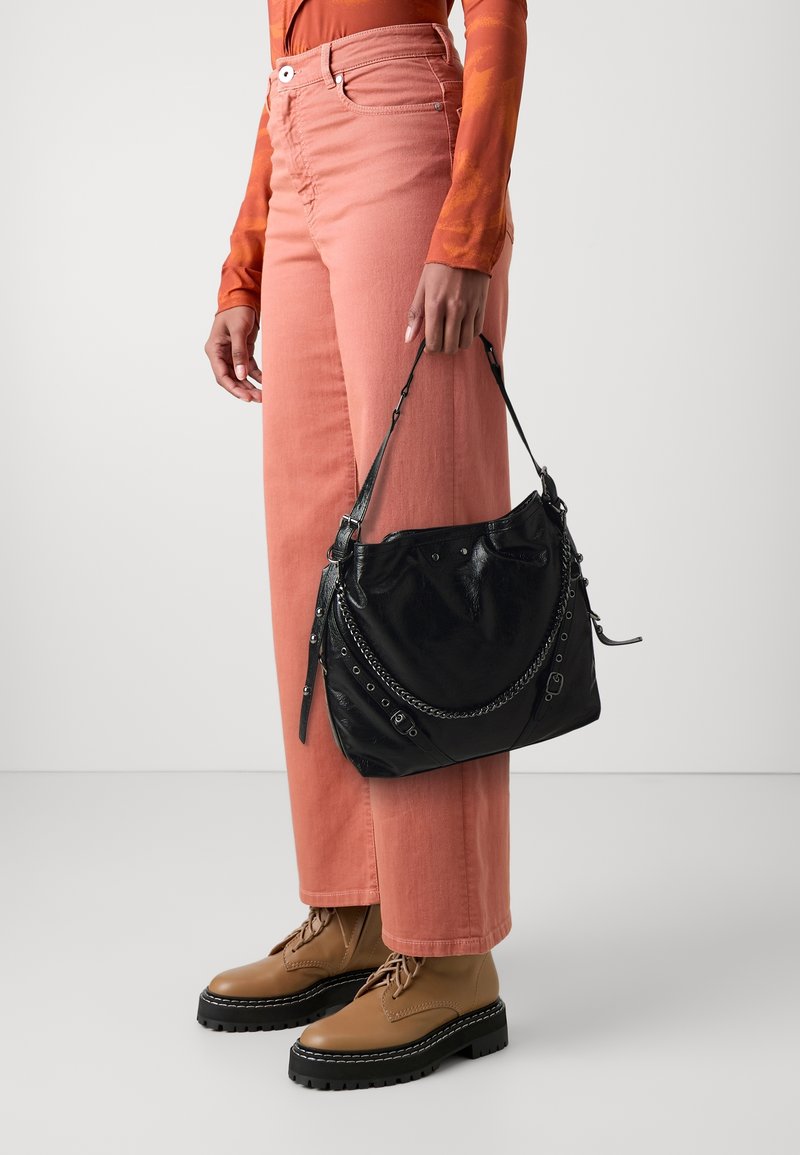 Personne portant un haut à manches longues orange, un pantalon rose saumon et des bottes lacées beige, tenant un sac à main en cuir noir avec des détails chaînes et boucle.