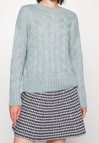 Ljusgrön kabelstickad tröja med ribbad nederkant och ärmslut, tillsammans med en svart- och vitfältmönstrad kjol. Båda plaggen har en texturerad yta.