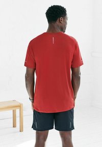 Rotes Sport-T-Shirt mit kurzen Ärmeln, glatter Textur, reflektierender Akzent auf dem Rücken, kombiniert mit schwarzen Shorts mit kleinen seitlichen Schlitzen.